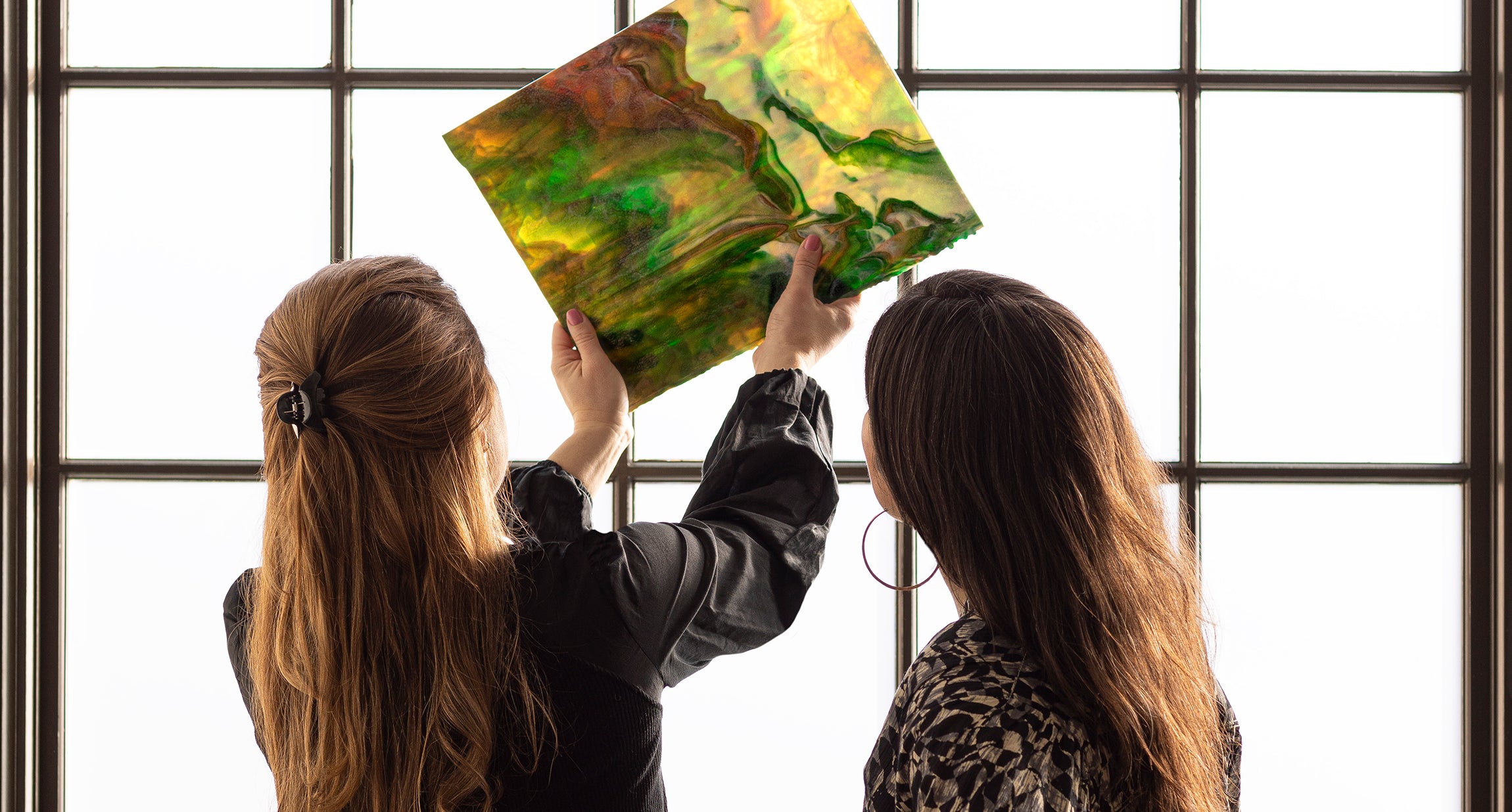 Two women standing in front of paned window-one holding square of window film.