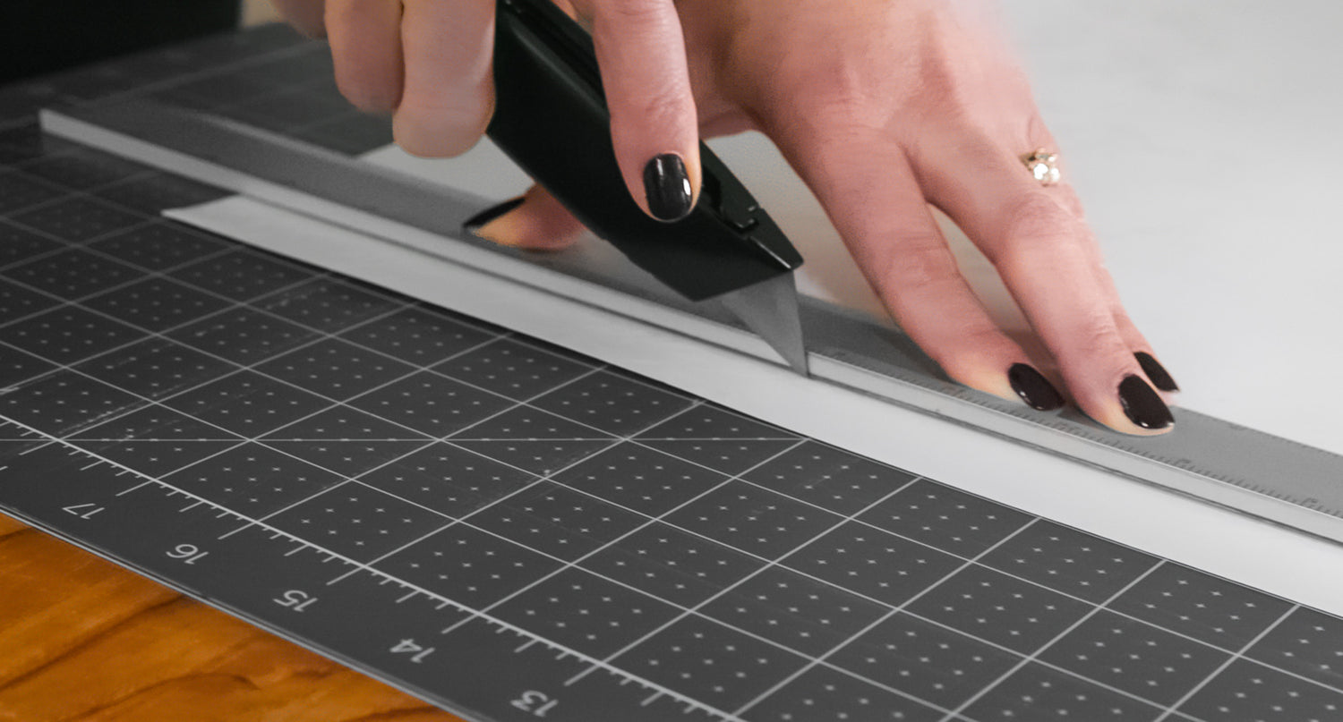View of hand using a utility knife to cut window film that is layed out on a cutting mat.