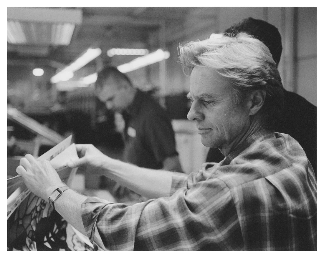 A monochrome image of a middle-aged man in a rolled-up sleeves plaid shirt examining sheets of window film, with men in workshirts in the background, under fluorescent ceiling lighting.