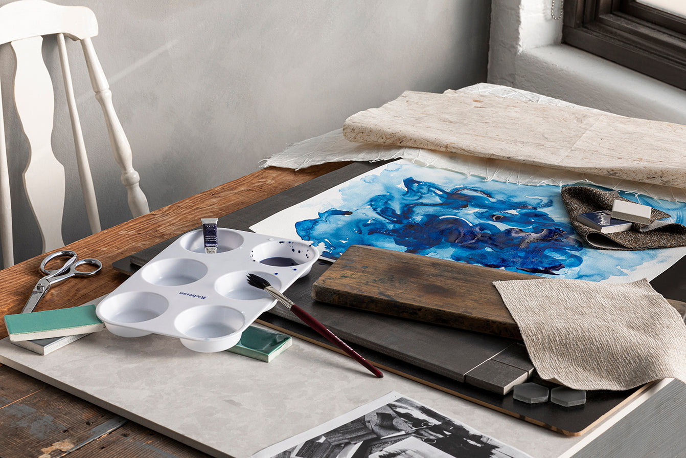 Desk spread with watercolor supplies and abstract watercolor painting in blue.