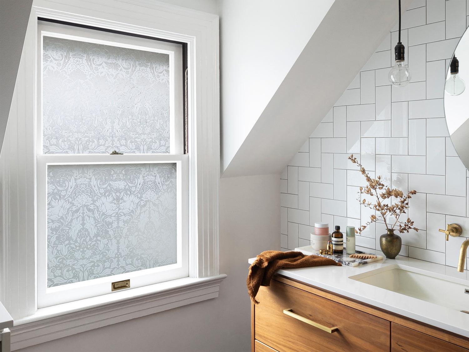 Chic bathroom interior with white walls, marble countertop, wooden cabinets, and a slanted dormer with an inset window featuring Artscape's Windsor window film with a classic floral design, enhancing privacy and style.