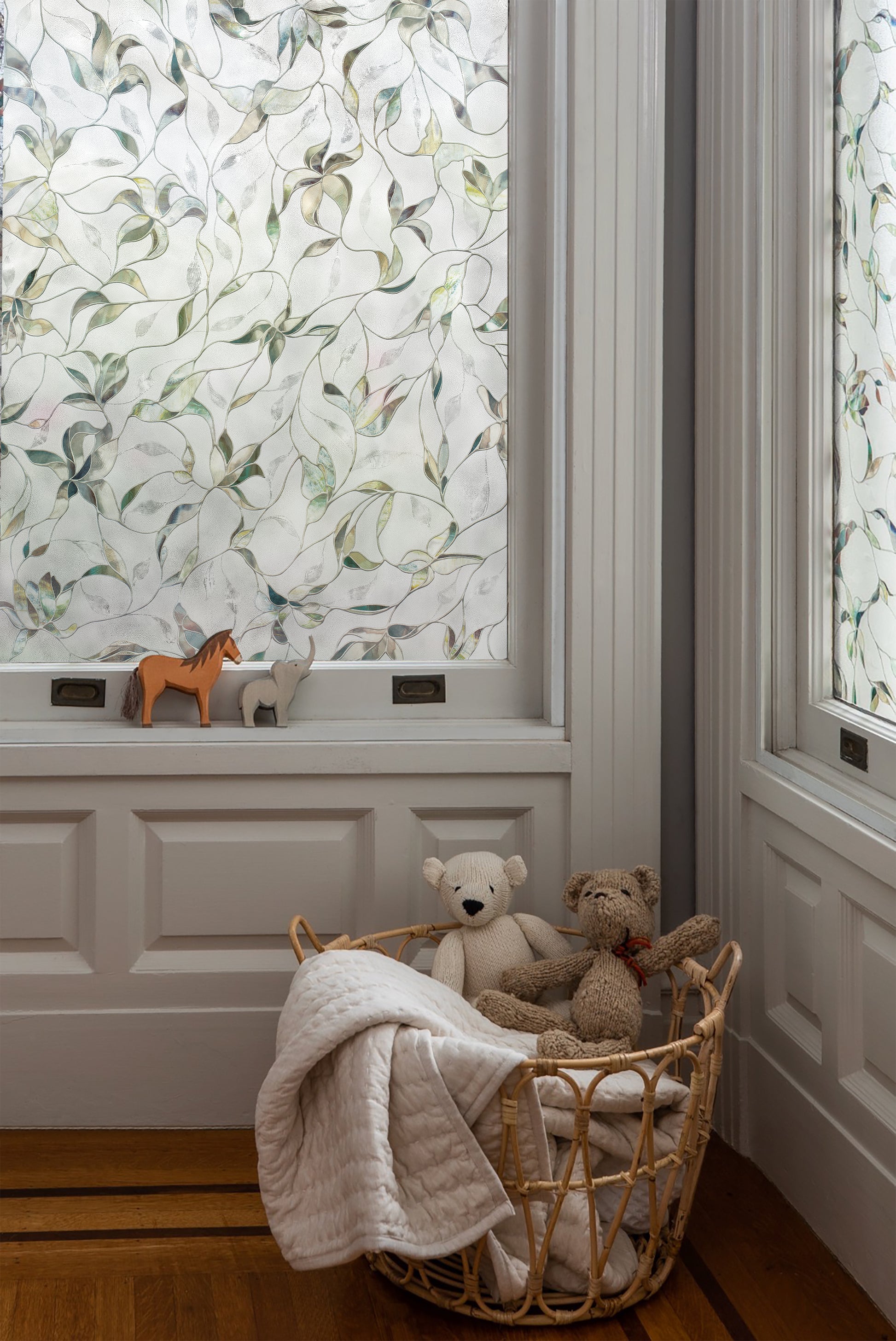Child's room with light grey walls and a classic, upscale feel, featuring a wicker basket filled with stuffed animals and blankets, toy wooden horses on the sill, and both windows adorned with Artscape's Flight window film for a touch of elegance.