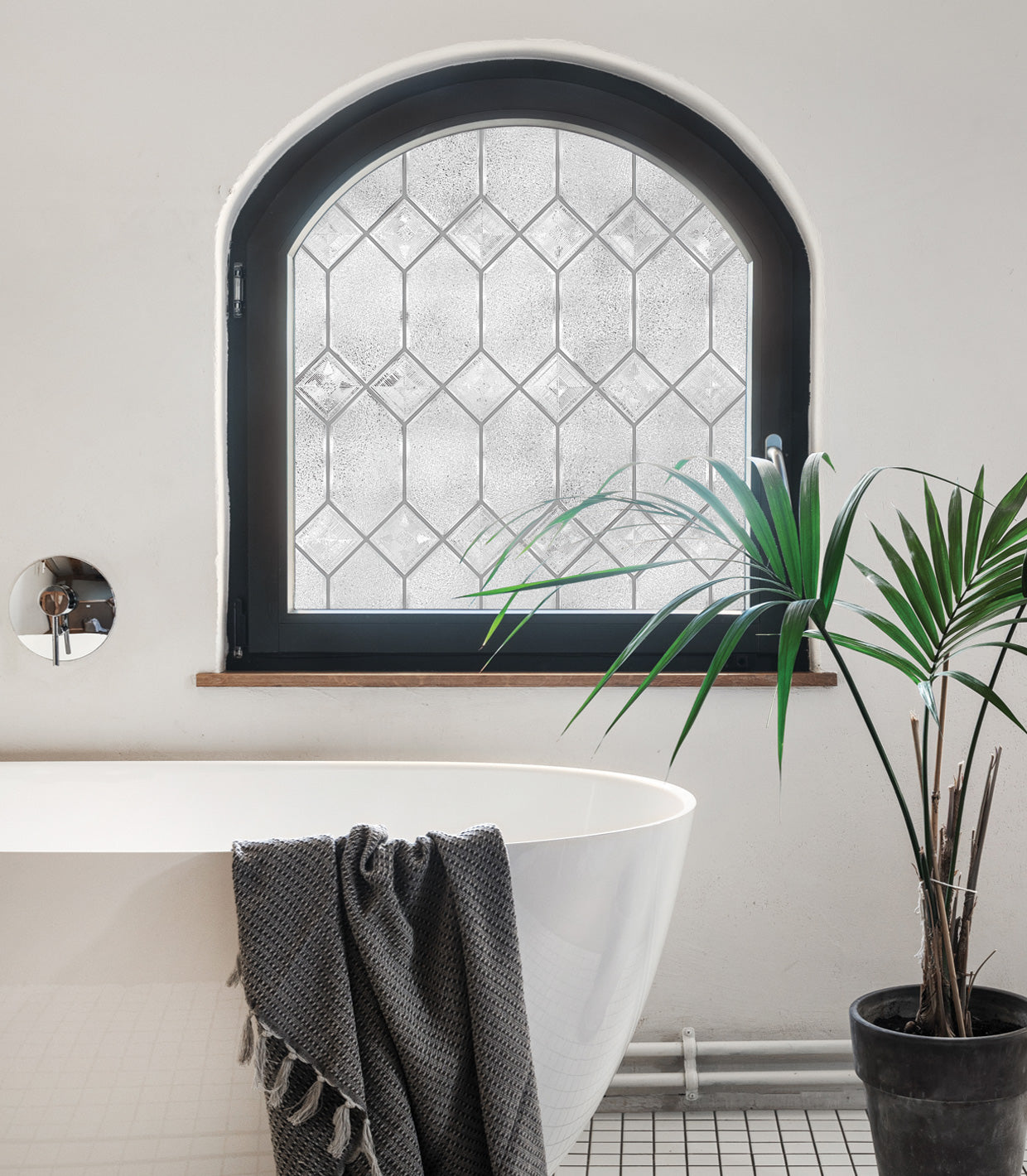 Modern, elegant bathroom featuring half of a white porcelain bathtub, tiled floor, and a potted palm plant. The room includes a window with a black frame and wooden sill, topped with a curved arch, enhanced with Artscape's Old English window film for classic appeal and privacy.