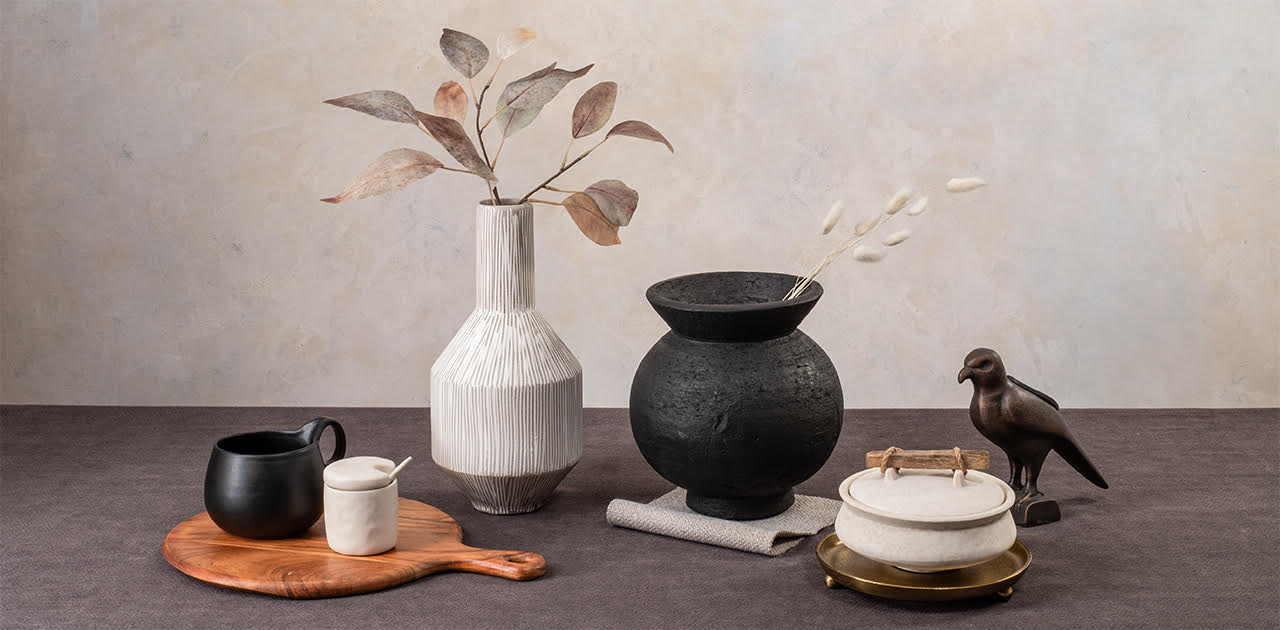 Minimalist still life on a dual-tone surface with assorted objects, including a matte jug, a white jar on a wooden board, a white vase with eucalyptus, a black pot with pampas grass, a marble container on a gold stand, and a dark bird figurine against a neutral wall.