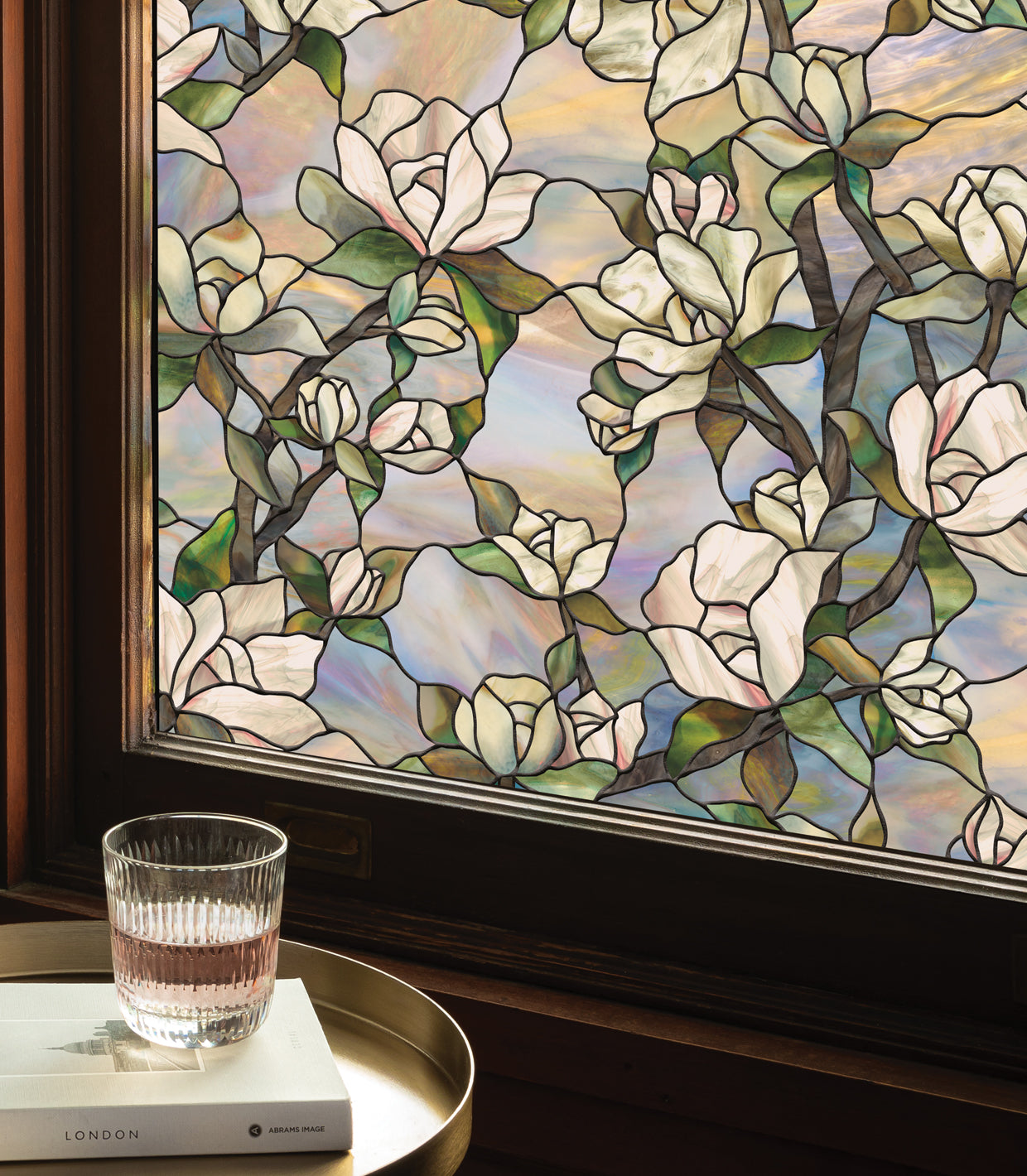 A drink in a glass placed on a book, which rests on a gold-colored metallic circular serving tray, set against a window with a rich, dark wood frame. The window is adorned with Artscape's Star Magnolia window film, a floral pattern that mimics stained glass, enhancing the room's aesthetic.