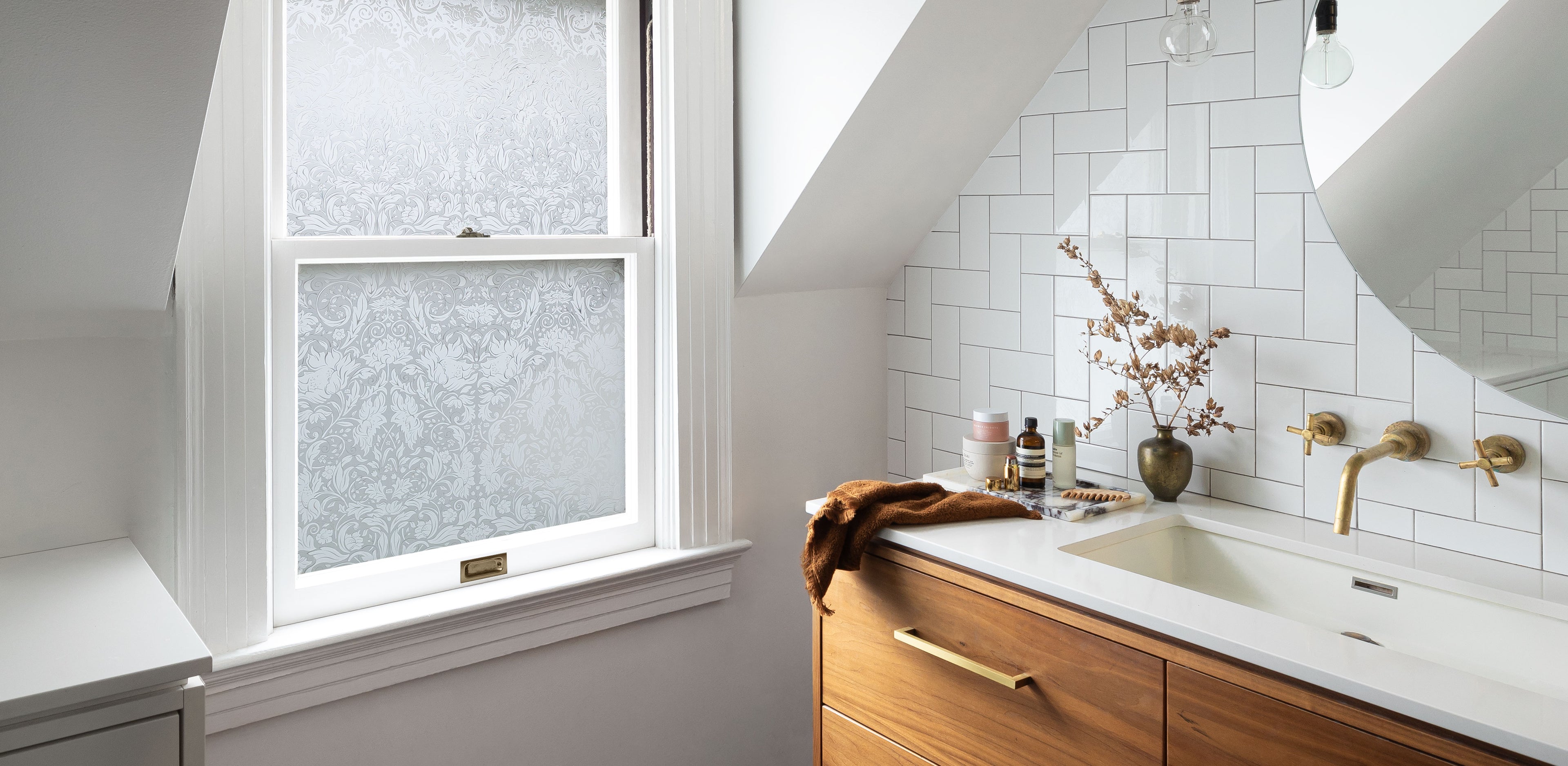 Chic bathroom interior with white walls, marble countertop, wooden cabinets, and a slanted dormer with an inset window featuring Artscape's Windsor window film with a classic floral design, enhancing privacy and style.